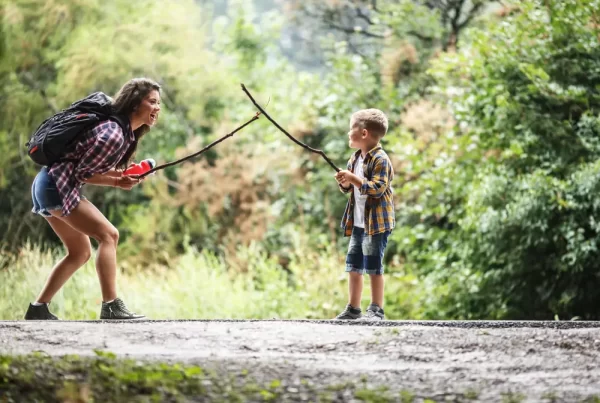 Mother and young boy playing imaginative stick sword game outdoors, demonstrating guided rough-and-tumble play in nature-based homeschool program.