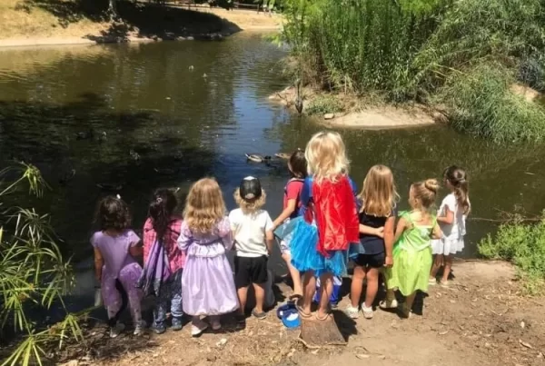 Children gathered at the edge of a pond during outdoor nature play, exploring the environment through hands-on, mud-friendly learning