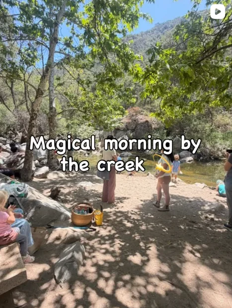 Children and families gathered beside a sunny creek surrounded by trees and mountains during a Nature Scouts Collective outdoor adventure, with text overlay reading ‘Magical morning by the creek.