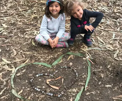 nature-scouts-children-nature-play Two young children sit outdoors under trees beside a heart made from leaves, bark, and natural materials, showing the joy and creativity of nature-based play.