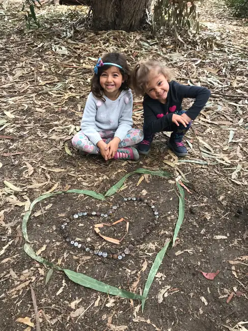 nature-scouts-children-nature-play Two young children sit outdoors under trees beside a heart made from leaves, bark, and natural materials, showing the joy and creativity of nature-based play.