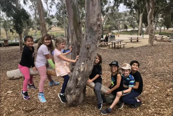 nature-scouts-children-outdoors Children from Nature Scouts Collective gather and play around a tree during an outdoor learning activity in a wooded park.